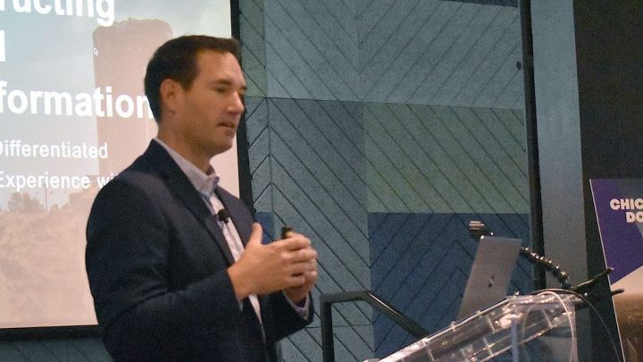 A man stands at a podium, speaking about how AI is transforming customer experience in construction equipment tracking.