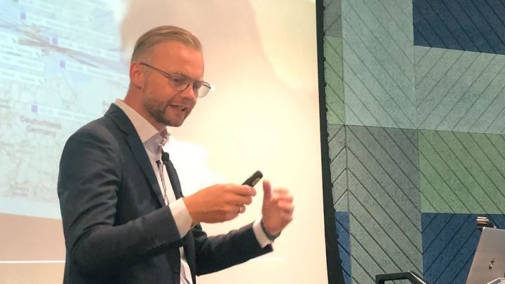 A man in a suit and glasses presents on construction’s future, with a map onscreen; relates to connected jobsite.