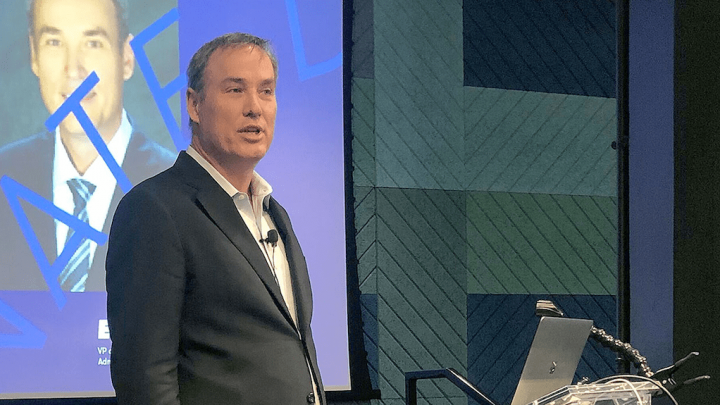 A man in a suit presents on reducing downtime for better customer experience, in a modern conference room with construction IoT focus.
