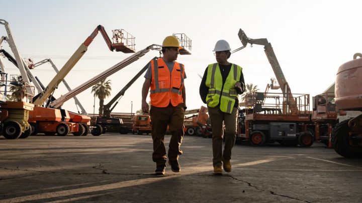 Two workers in safety gear walk and talk in a lot of aerial lifts and smart machinery, highlighting a connected jobsite.