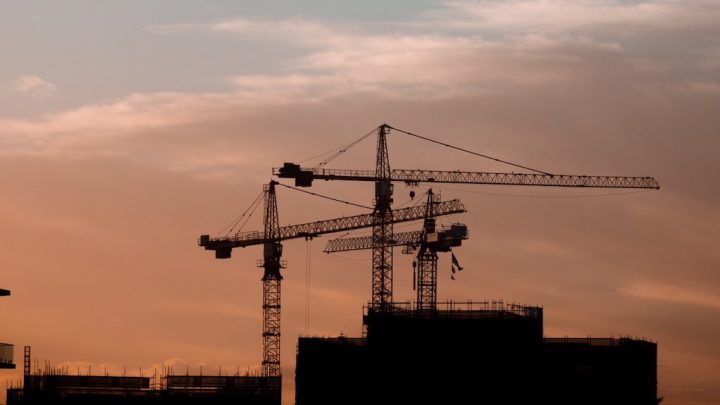 Three construction cranes and unfinished buildings silhouetted against an orange-pink sunset sky, illustrating a connected jobsite.