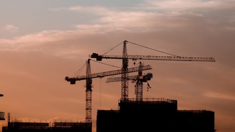 Three construction cranes and unfinished buildings silhouetted against an orange-pink sunset sky, illustrating a connected jobsite.