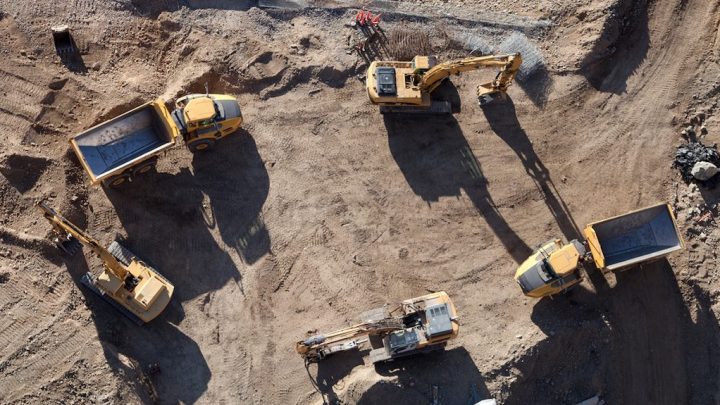 Aerial view of a construction site with four yellow trucks and two excavators; dirt, tire tracks, and equipment show construction IoT in use.