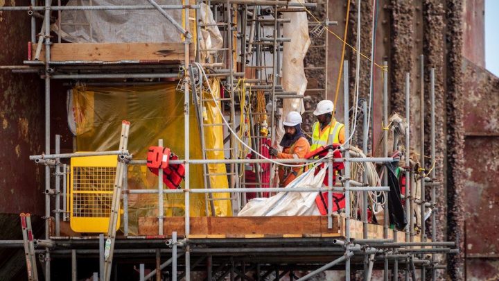 Two construction workers in safety gear review data on scaffolding at a connected jobsite with a partially covered structure behind them.