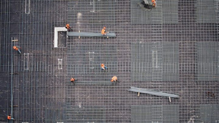 Aerial view of workers in orange vests arranging rebar grids on a concrete foundation using digital construction IoT guidance.