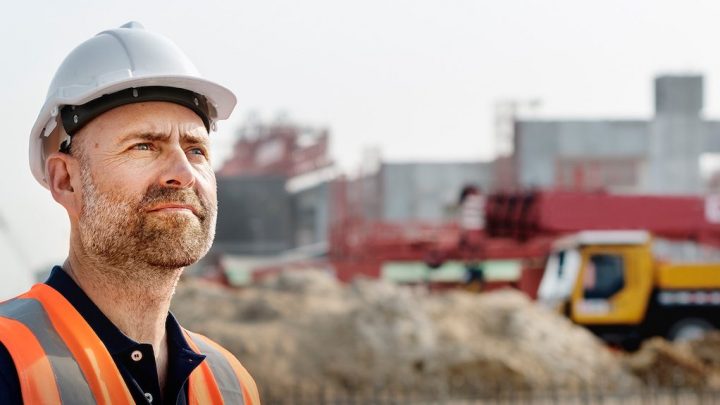 A construction worker in a hard hat and safety vest surveys a site with machinery and structures, highlighting construction equipment tracking.