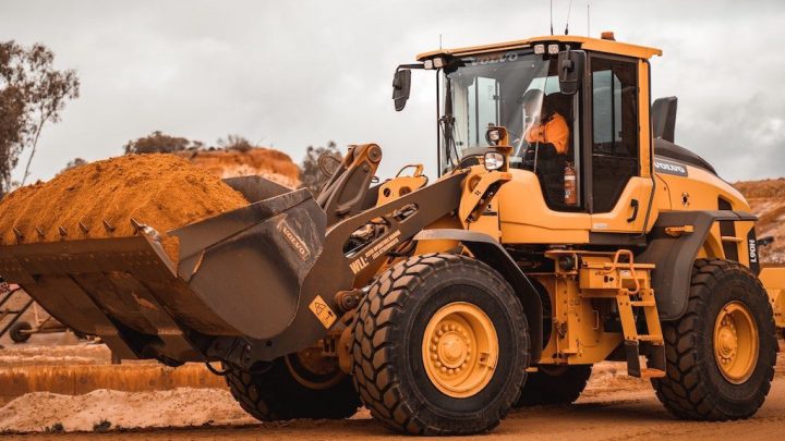 A yellow front-loader with a full bucket of dirt is parked on a jobsite, operator inside, showing construction equipment tracking.
