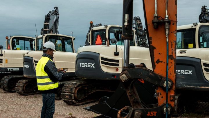 A worker in a yellow vest inspects white TEREX excavators parked outdoors on a cloudy day, as part of fleet management.
