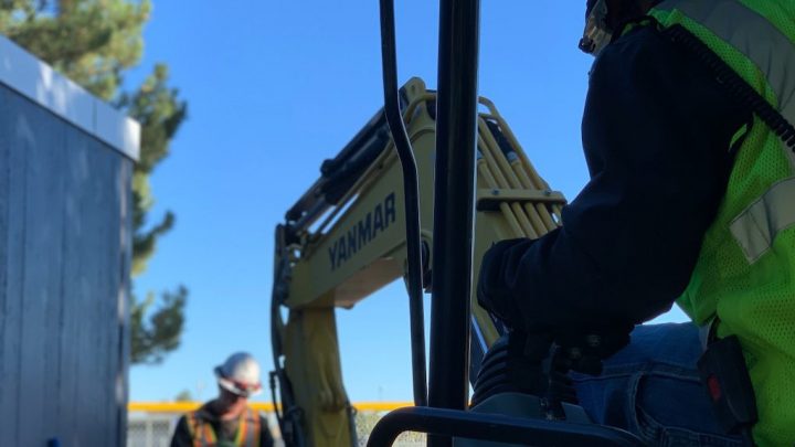 A worker uses a Yanmar excavator as another stands nearby; tools management software keeps this connected jobsite organized.
