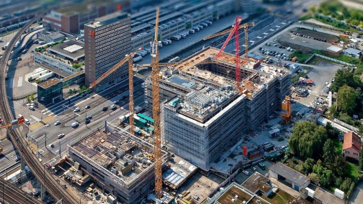 Aerial view of an urban construction site with cranes and scaffolding, showing construction IoT in use among nearby roads and buildings.