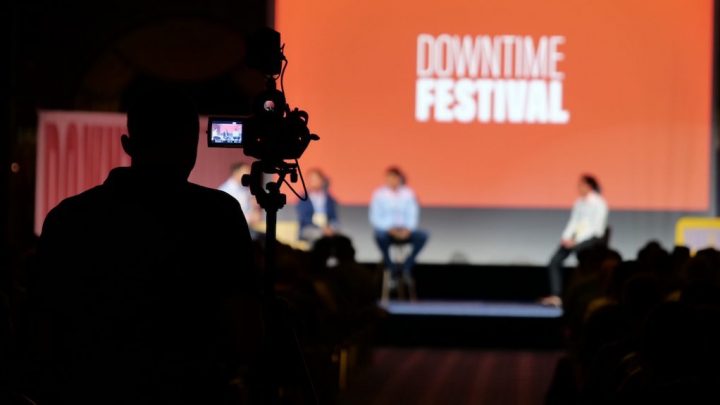 A silhouette of a cameraman films a panel at Downtime Festival, with “product portal” onscreen and a dimly lit audience in front, highlighting connected jobsite.