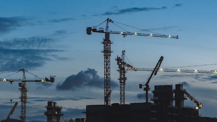 Construction cranes tower over a building site at dusk, silhouetted against a blue sky—showcasing connected jobsite coordination.