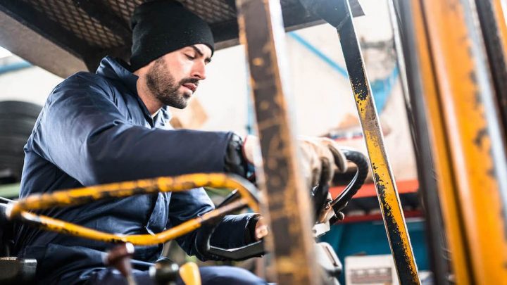 A man in a beanie and gloves operates a forklift in an industrial warehouse, focused on controls, showing good fleet management.