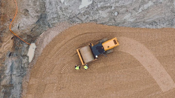 Aerial view of a construction site with a yellow bulldozer leveling soil to ISO standard; two workers and tire tracks visible, construction.