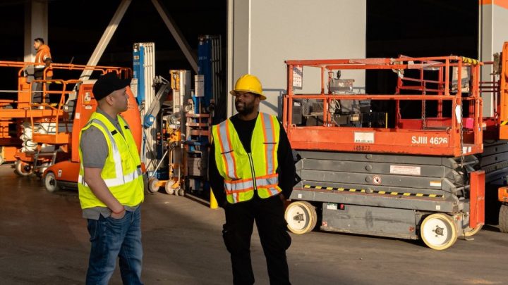 Two workers in safety gear discuss fleet management in a sunlit warehouse with orange scissor lifts and equipment in the background.