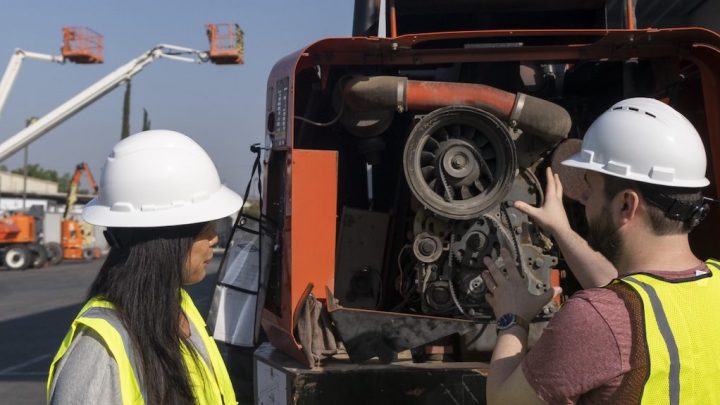 Two workers in safety gear inspect a large machine’s engine outdoors, with boom lifts behind them, emphasizing fleet management.