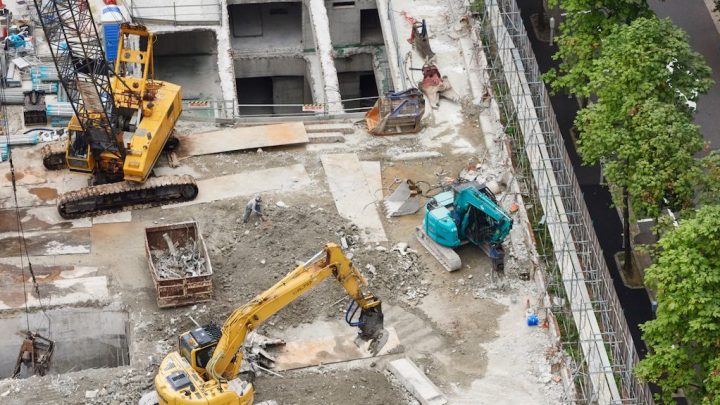 Aerial view of a construction site with cranes, excavators, and materials near trees and a road, illustrating fleet management.