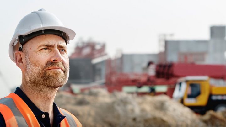 A man in a white hard hat and orange vest stands at a construction site, looking thoughtful. Blurred equipment hints at fleet management.