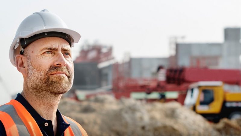 A man in a white hard hat and orange vest stands at a construction site, looking thoughtful. Blurred equipment hints at fleet management.