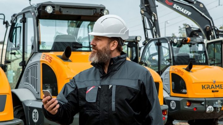 A man in a white hard hat uses a smartphone outdoors, with yellow construction vehicles and excavators in the background, showing fleet management.
