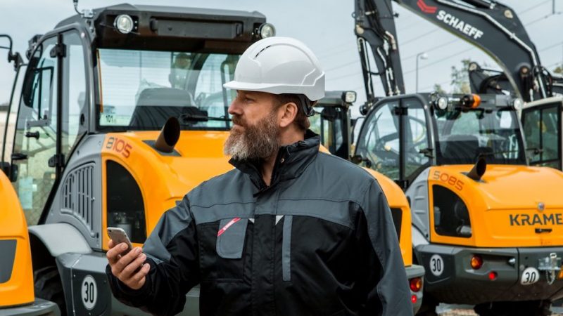 A man in a white hard hat uses a smartphone outdoors, with yellow construction vehicles and excavators in the background, showing fleet management.
