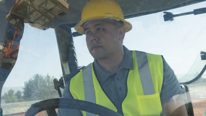 A man in a yellow hard hat and safety vest sits in a construction vehicle cab, looking ahead, illustrating telematics in fleet management.