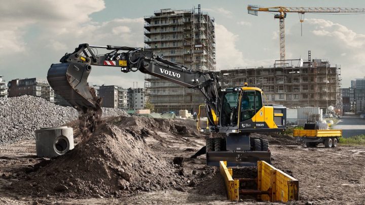 A Volvo excavator with GPS works at a construction site, near a multi-story build and crane, showing connected jobsite activity.
