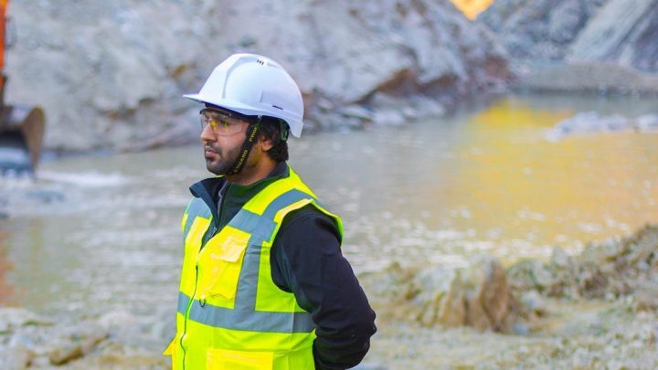 A person in safety gear stands at a muddy construction site near water, with excavation equipment and construction IoT tools behind them.