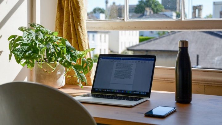 A laptop on a wooden desk by a window with a plant, water bottle, and smartphone—remote work and connected jobsite in sunlight.