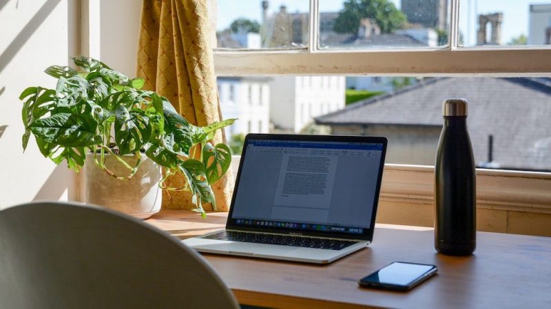 A laptop on a wooden desk by a window with a plant, water bottle, and smartphone—remote work and connected jobsite in sunlight.