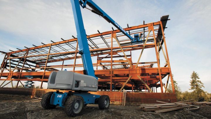 A blue boom lift with telematics is on rough ground near a steel-framed building under construction at a connected jobsite.