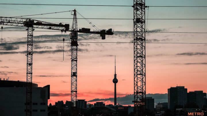 Construction cranes against a pink-blue sunset with city skyline and power lines, suggesting a connected jobsite.