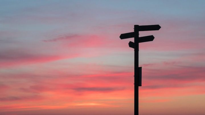A silhouetted signpost with arrow signs stands against a colorful sunset sky, often seen near construction equipment tracking sites.