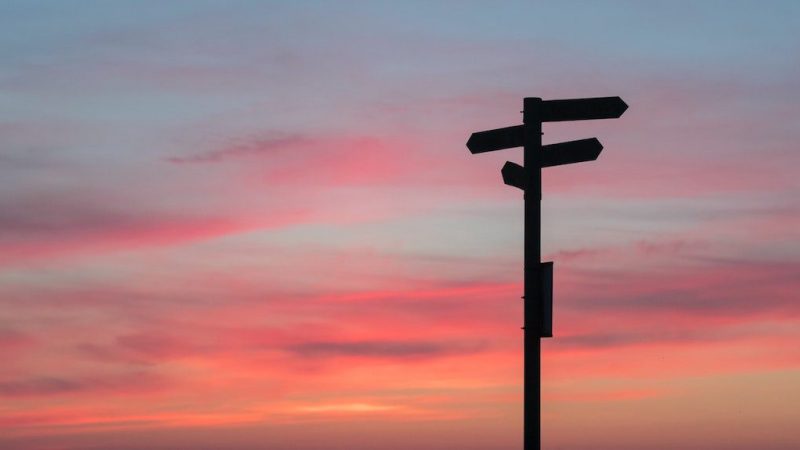 A silhouetted signpost with arrow signs stands against a colorful sunset sky, often seen near construction equipment tracking sites.