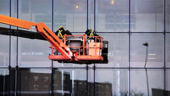 Two workers in safety gear use an orange boom lift to clean glass building windows, with city reflections and construction equipment tracking.