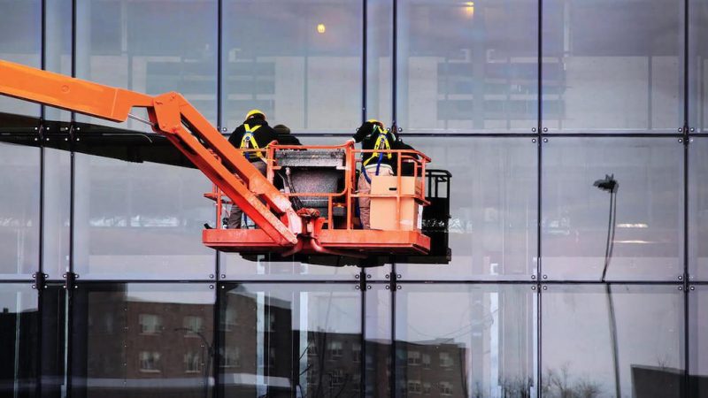 Two workers in safety gear use an orange boom lift to clean glass building windows, with city reflections and construction equipment tracking.