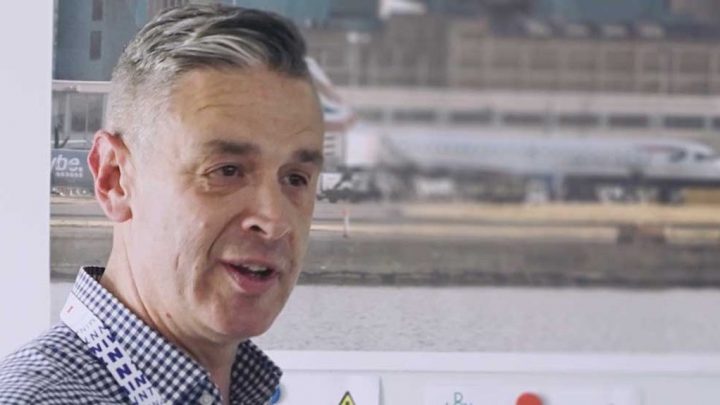 A man with short gray hair in a checkered shirt and lanyard speaks by an airport runway, discussing construction fleet management.