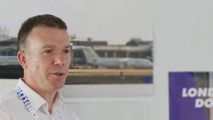 A man in a white shirt and lanyard stands indoors with a blurred airport and predictive fleet management activity in the background.