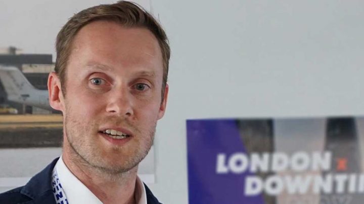 A man in a suit stands indoors by a blurred "LONDON x DOWNTIME" sign and a photo symbolizing construction productivity.