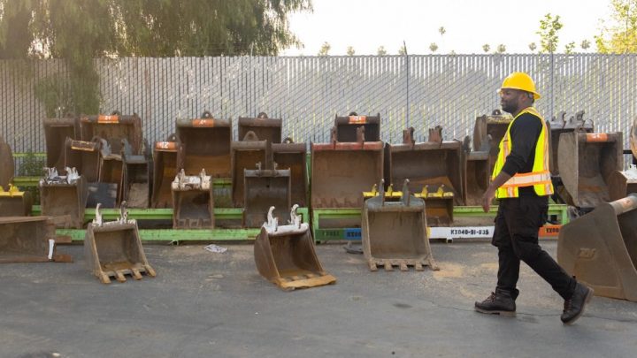 A man in a yellow hard hat and vest walks by rows of excavator buckets outdoors, highlighting fleet management.