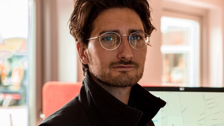 A man with wavy brown hair and glasses looks at the camera indoors, with fleet management software open on a computer behind him.