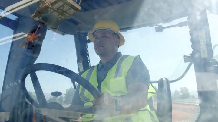 A man in a yellow hard hat drives construction equipment, seen through a reflective window—showcasing fleet management use.