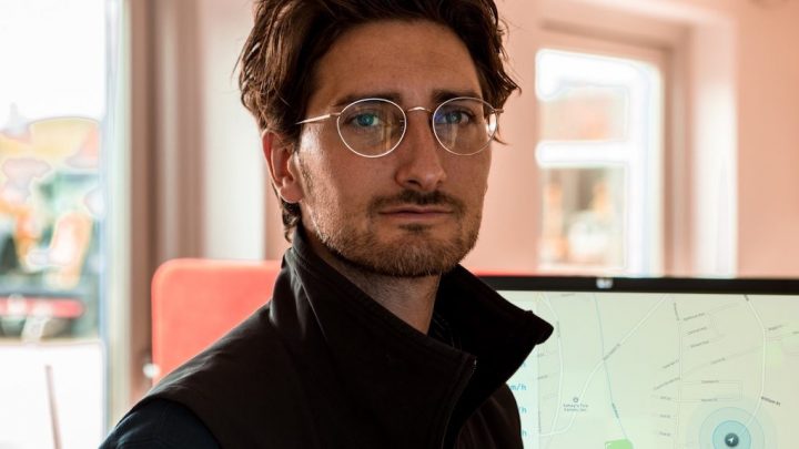 A man with glasses and brown hair looks at the camera; behind him, a monitor shows construction equipment tracking software and a map.