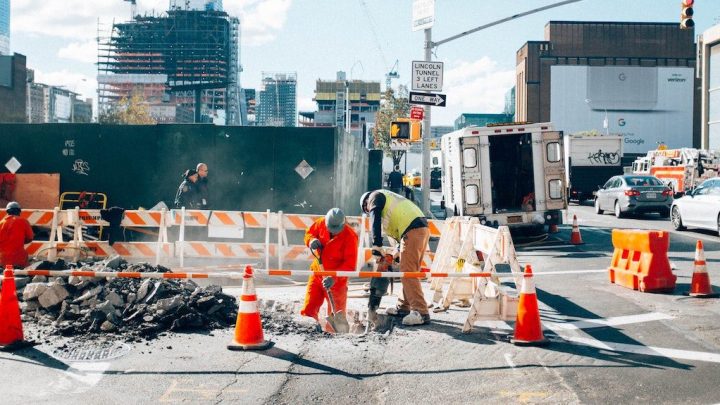 Construction workers repair a city street with cones and barriers, set against a cityscape, showing connected jobsite technology.