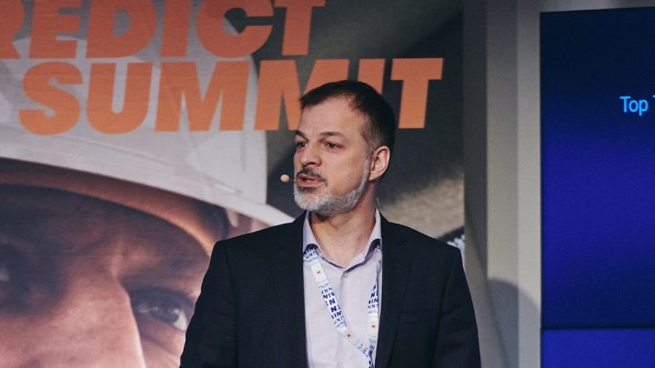 A man in a suit with a headset speaks at a conference with a PREDICT SUMMIT banner and screen, related to construction IoT.