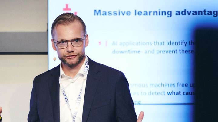 A man in a suit speaks at a conference on AI and machine learning, with a device in hand and a slide about construction IoT behind him.