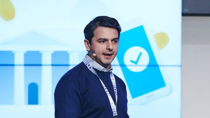A man speaks into a headset onstage at a conference, with a screen behind him showing a building and smartphone graphic for connected jobsite.