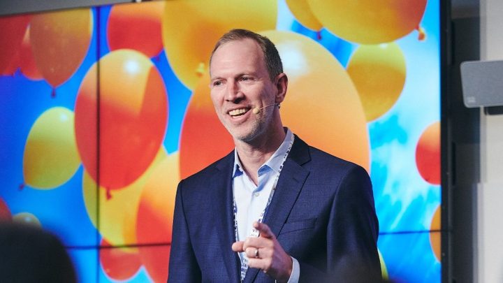 A man in a blue suit speaks on stage with a headset mic, smiling and gesturing, at a conference with a colorful balloon display.
