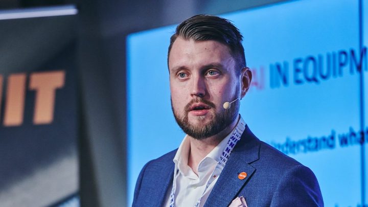 A bearded man in a blue suit with a headset speaks at a conference on construction IoT, standing before a screen with text.