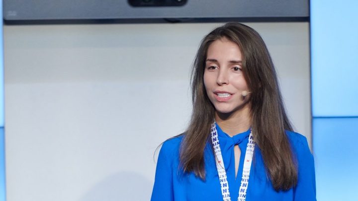 A woman with long brown hair in a blue shirt speaks on stage at a conference about connected jobsite solutions.